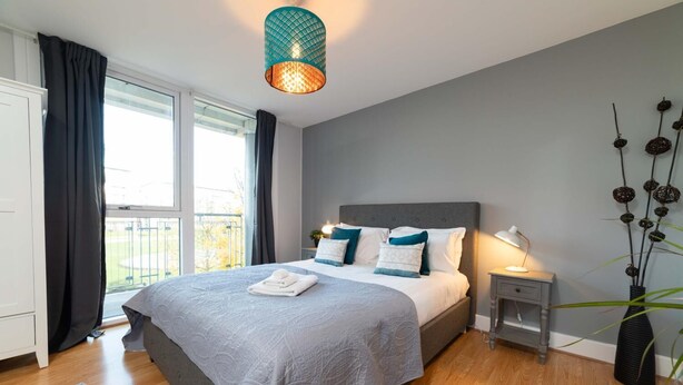 Une chambre moderne avec un grand lit, une table de chevet avec une lampe, une vue sur l’extérieur par une fenêtre et une plante décorative.