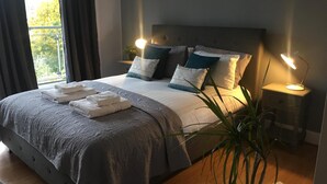 Un lit bien fait, avec une couette grise, des oreillers et des serviettes pliées. On y trouve deux tables de chevet avec des lampes, une grande fenêtre avec des rideaux et une plante en pot.