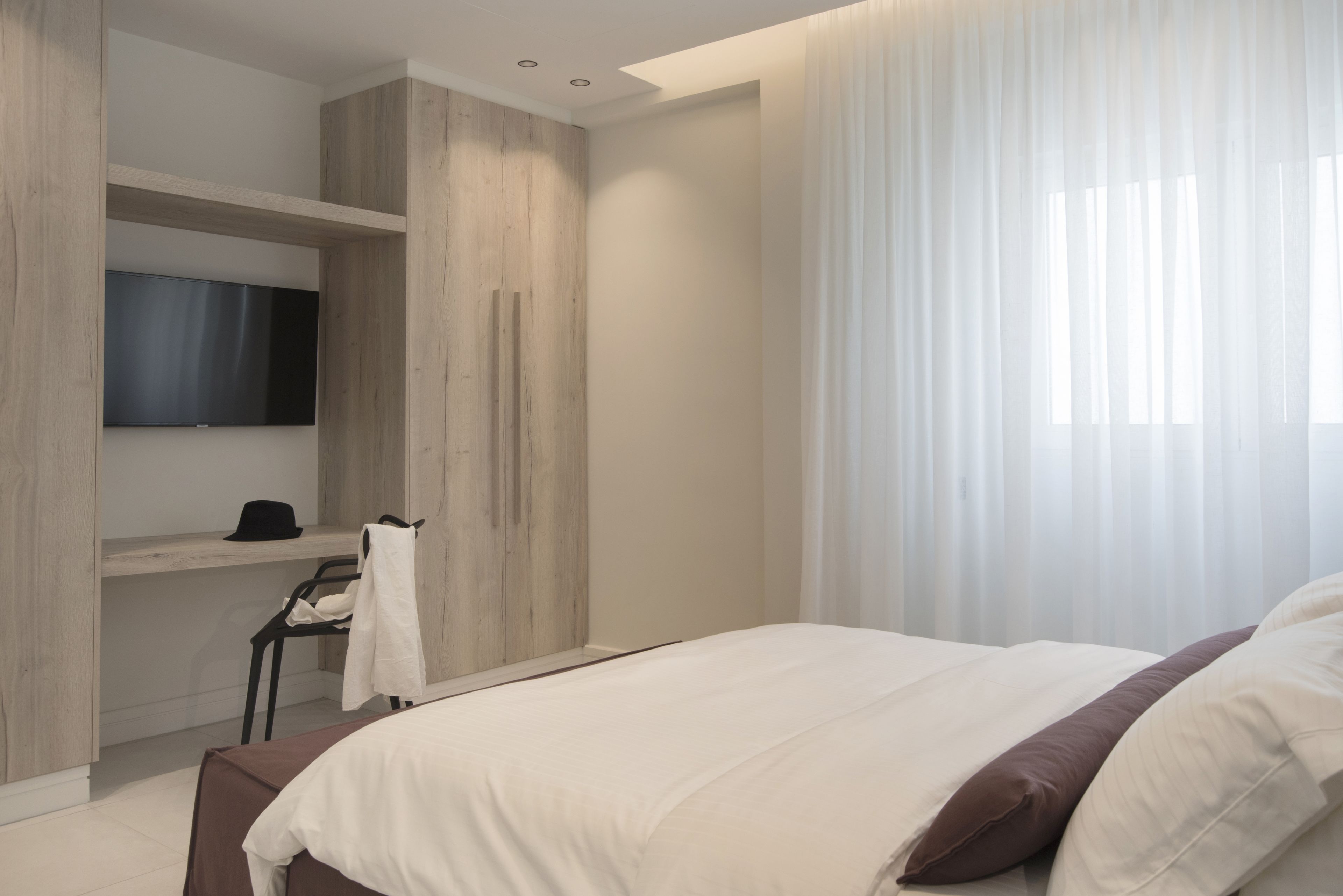 A modern bedroom with a large bed, a built-in TV, a wardrobe, and a window with sheer curtains.
