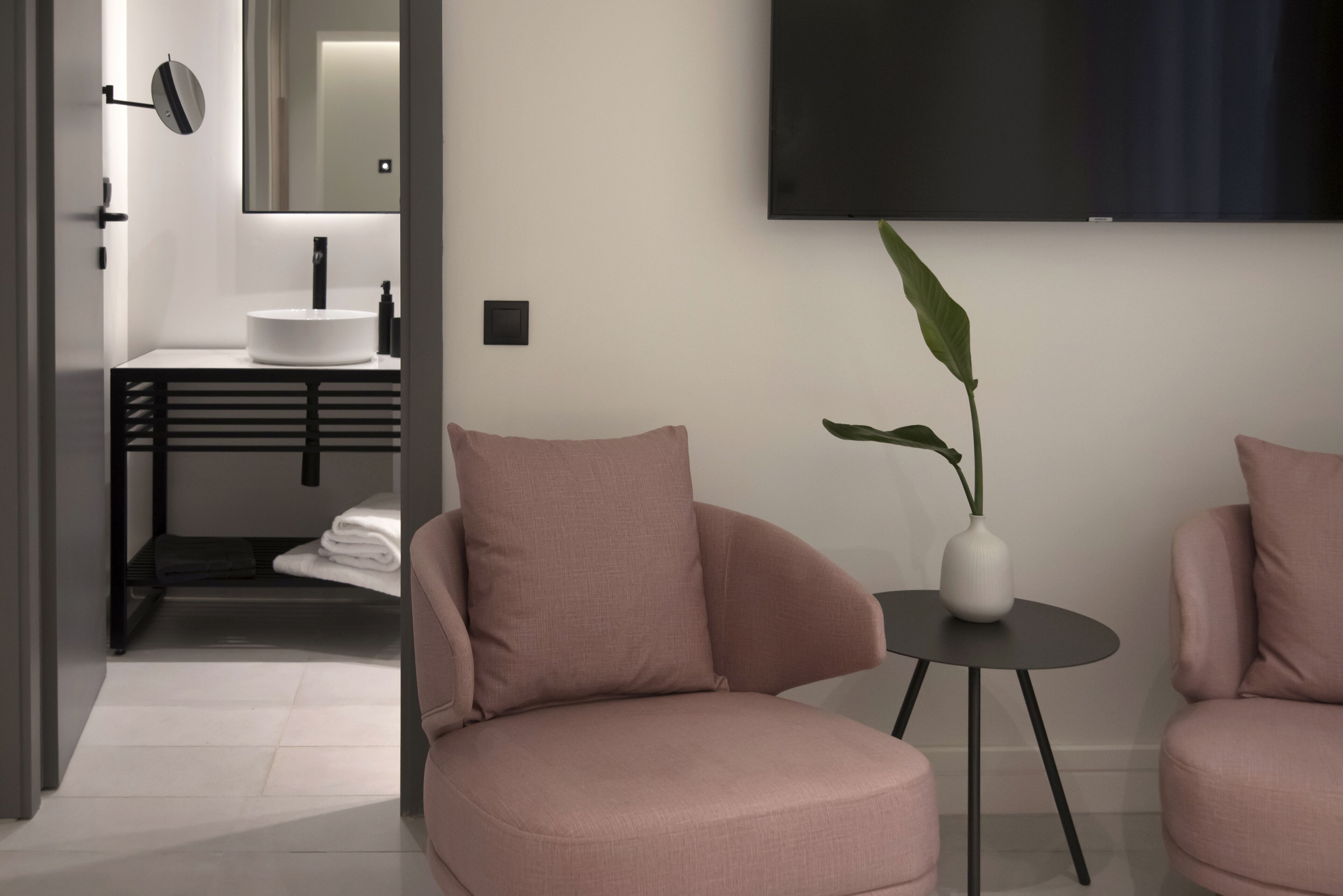 A modern bathroom with a white sink, black towel rack, and a mirror. There is a black wall-mounted TV above a round table with a white vase and a plant.