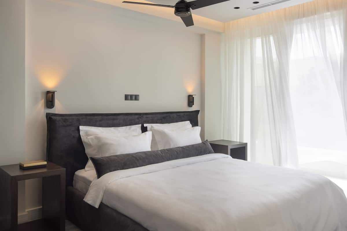 A modern hotel room with a large bed, two bedside tables, a ceiling fan, and a window with sheer curtains.
