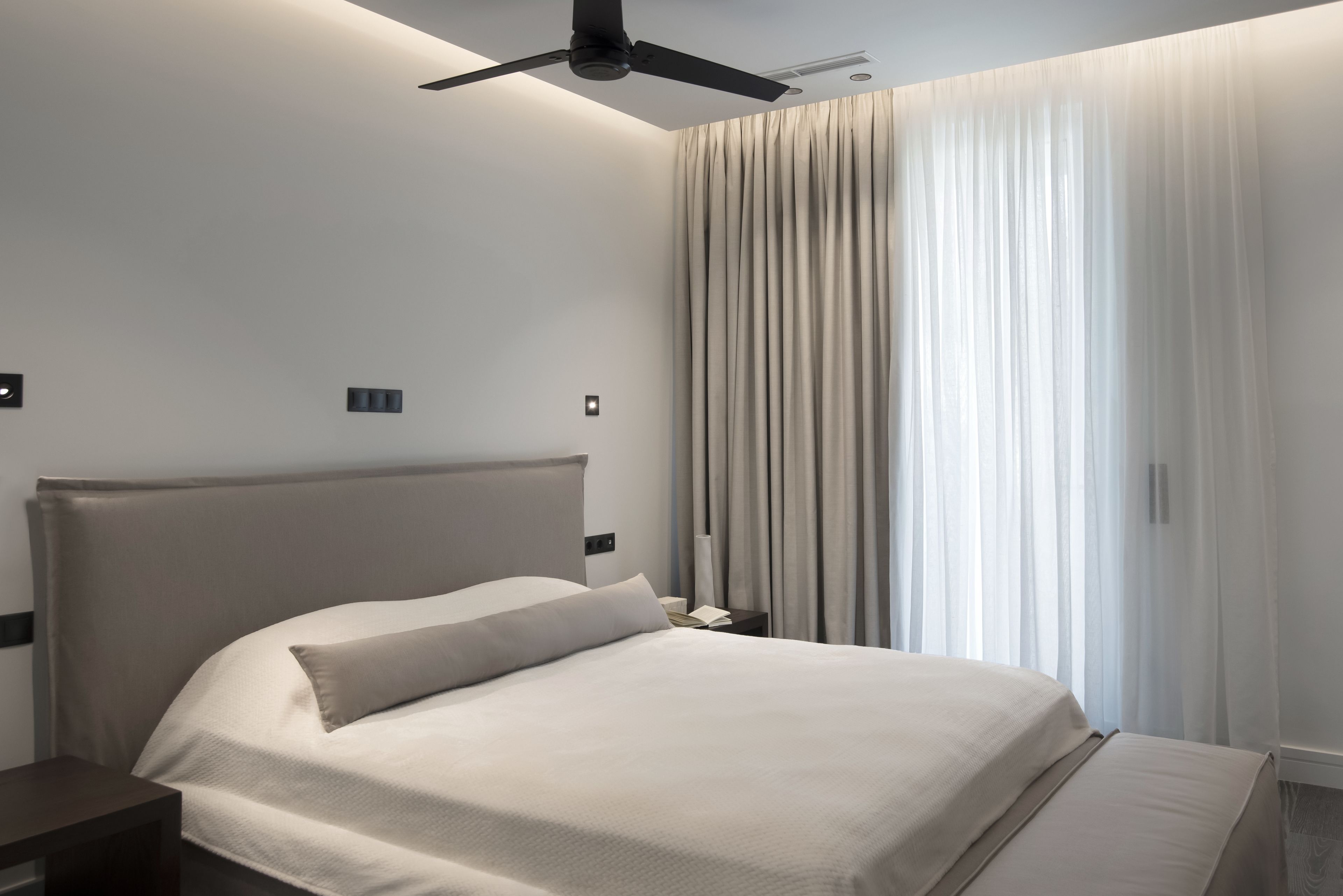 A modern hotel room with a large bed, a ceiling fan, and a view of the city through sheer curtains.