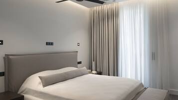 A modern hotel room with a large bed, a ceiling fan, and a view of the city through sheer curtains.