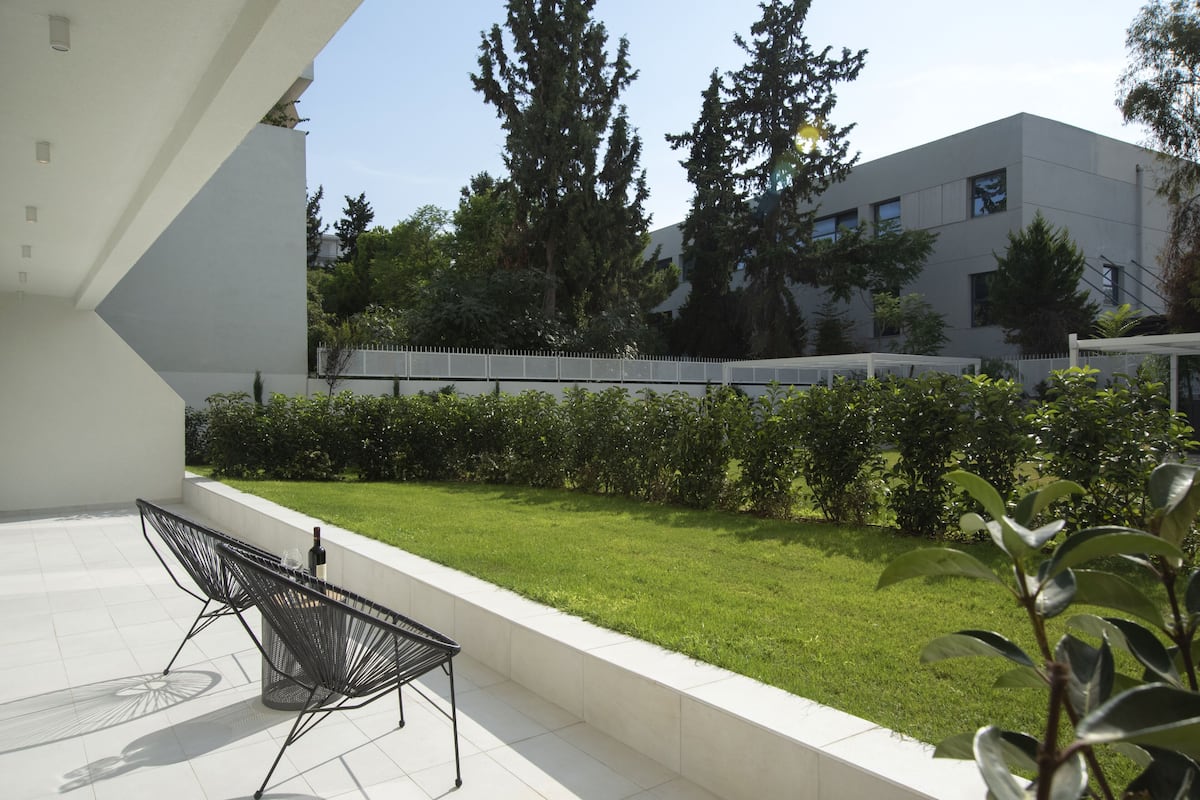 A modern outdoor patio with a metal table and chairs, a white wall, and a well-maintained lawn with trees and a fence in the background.