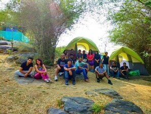 A group of people sitting and standing around two green tents in a natural setting with rocks and trees. - Hasiru Holidays (Anekal)