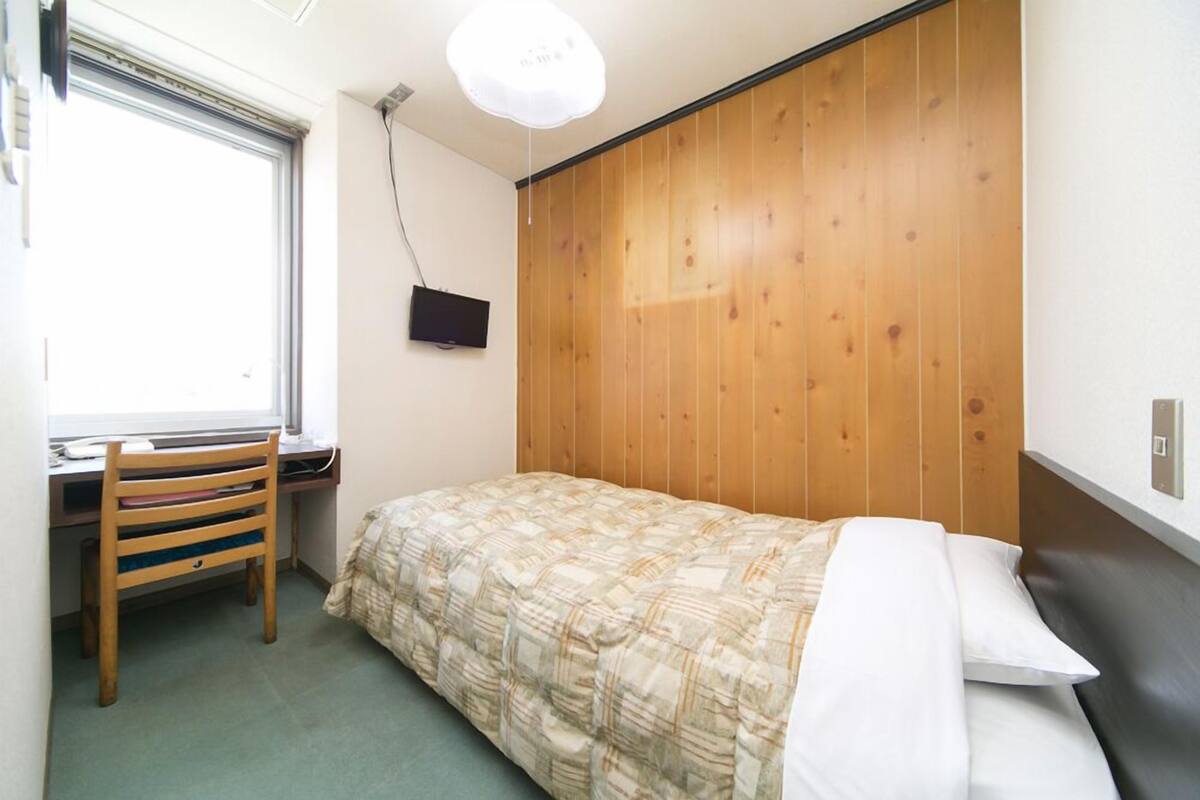 Un dormitorio pequeño y ordenado con cabecera de madera, una cama individual, un escritorio con silla y una ventana.