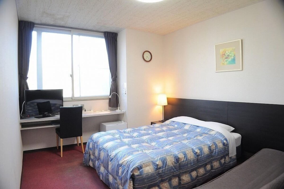 Una habitación de hotel con cama, escritorio, silla y ventana con cortinas.