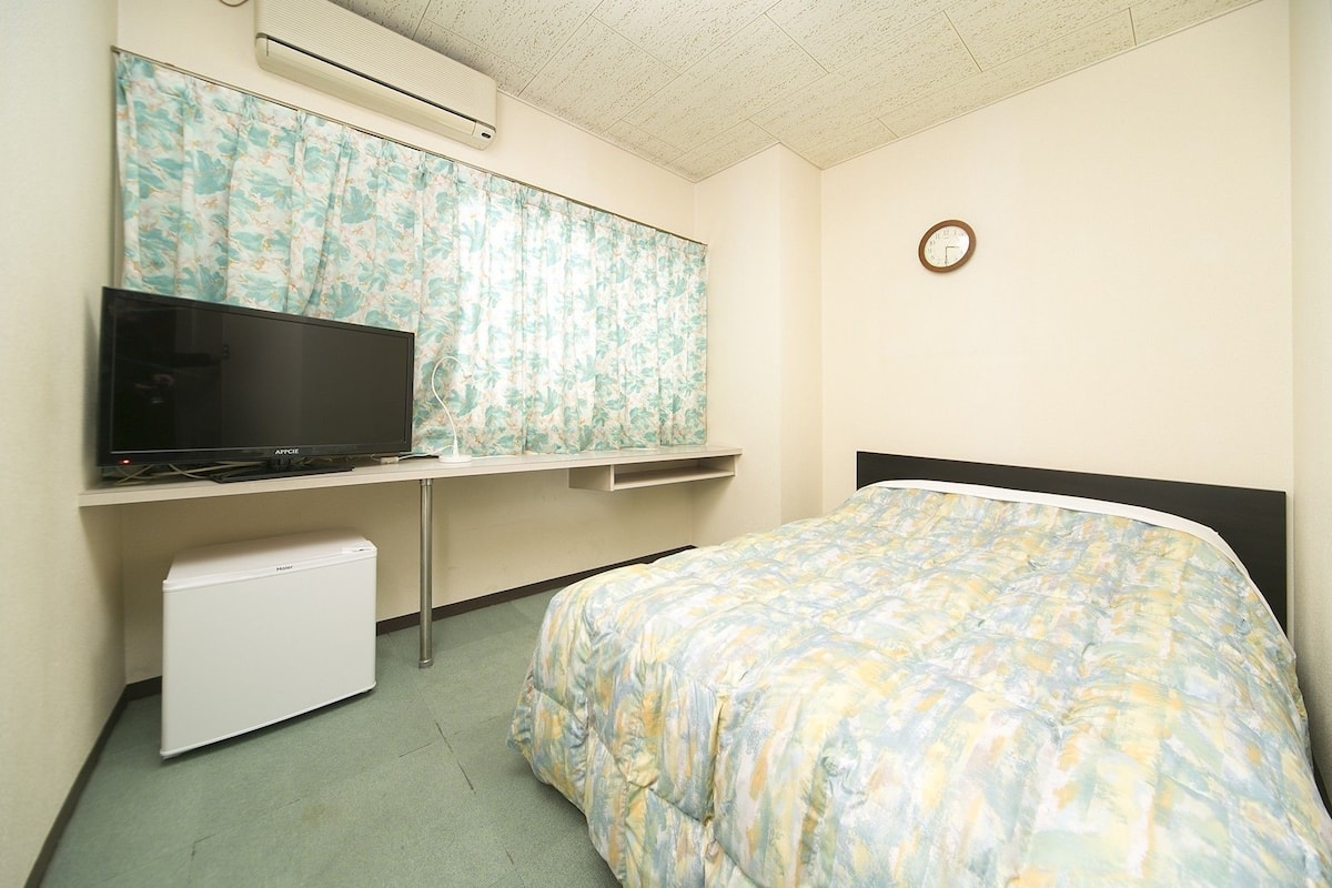 Una habitación pequeña y sencilla con una cama, un escritorio con televisor y un refrigerador pequeño.