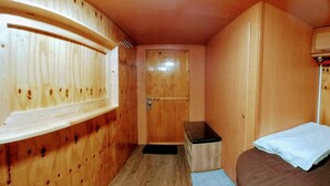 A small, compact room with wooden walls and a single door. There is a bed with a white blanket and a small table with a black top. - Samui Caravans Hill Camp (Koh Samui)