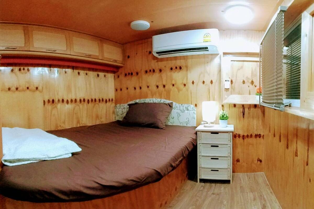 A wooden cabin room with a bed, a bedside table with a lamp, and an air conditioning unit.