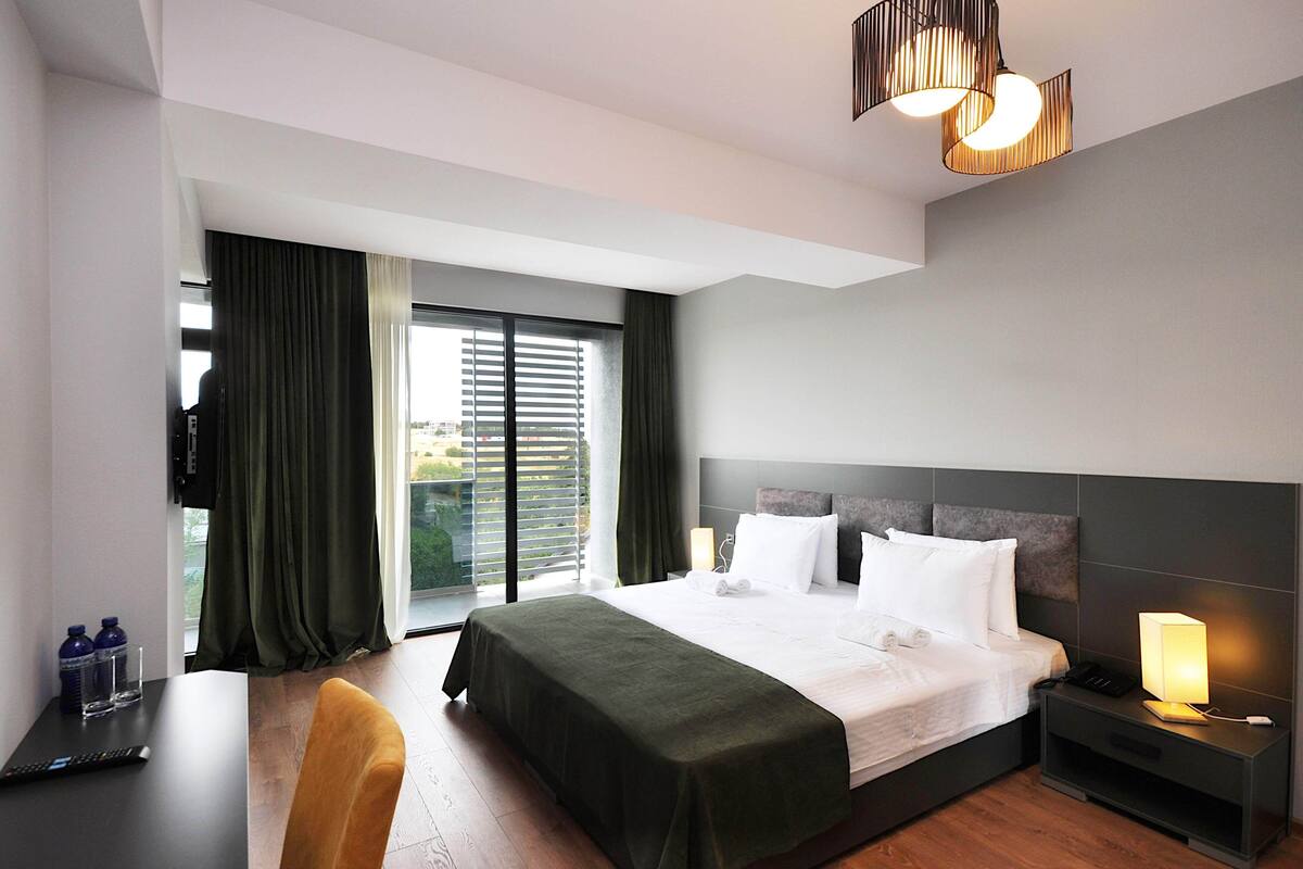 A modern hotel room with a large bed, a desk, a chair, a nightstand with a lamp, and a view of the outdoors through the window.