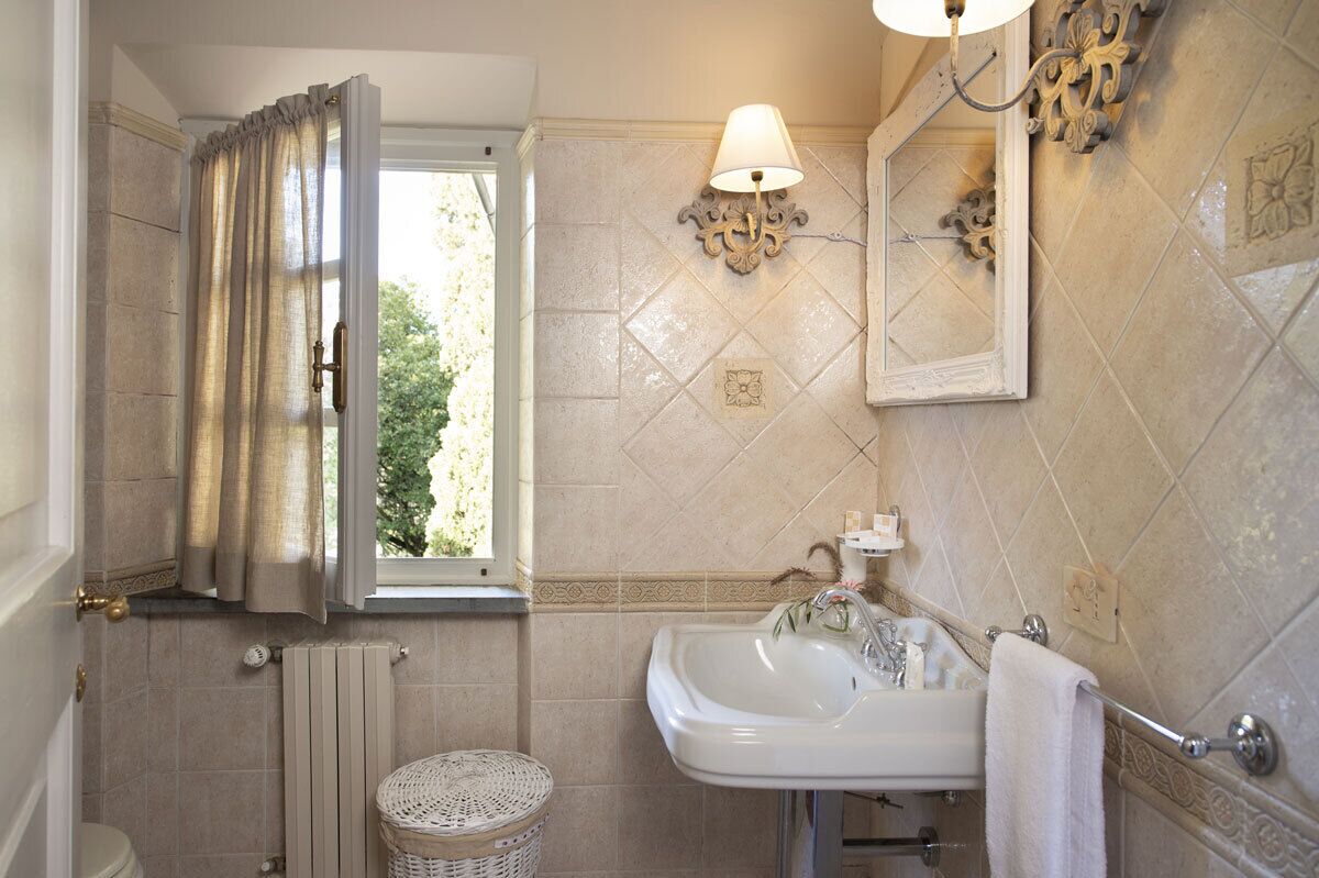 Une salle de bain avec un lavabo blanc, un miroir, un luminaire fixé au mur et une fenêtre avec des rideaux.