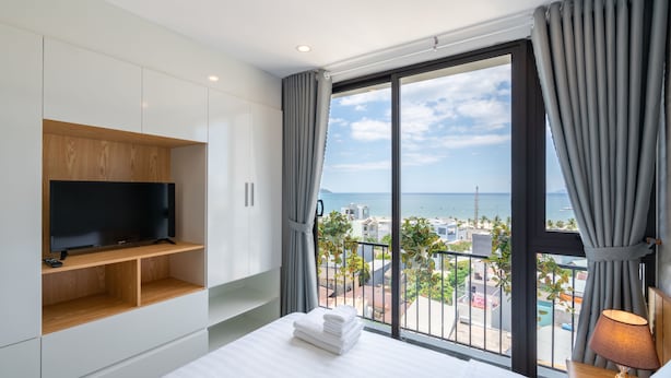 A modern hotel room with a large bed, a flat-screen TV on a wooden shelf, a balcony with a view of the sea, and a lamp on the bedside table. - Dandelion Apartment (Da Nang)