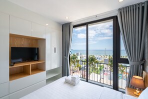 A modern hotel room with a large bed, a flat-screen TV on a wooden shelf, a balcony with a view of the sea, and a lamp on the bedside table. - Dandelion Apartment (Da Nang)