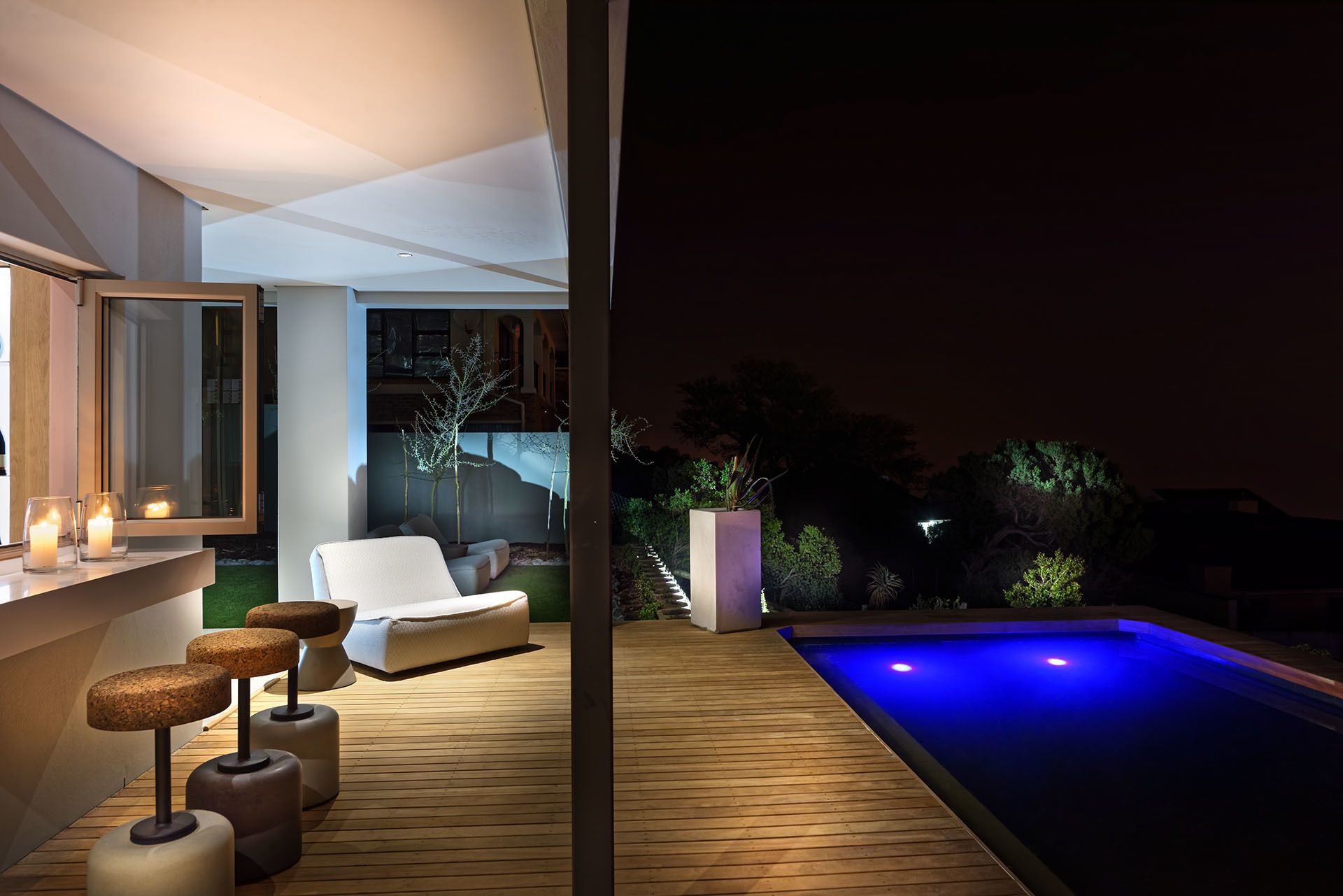 A modern outdoor pool area with a wooden deck, a lounge chair, and a small table with candles.