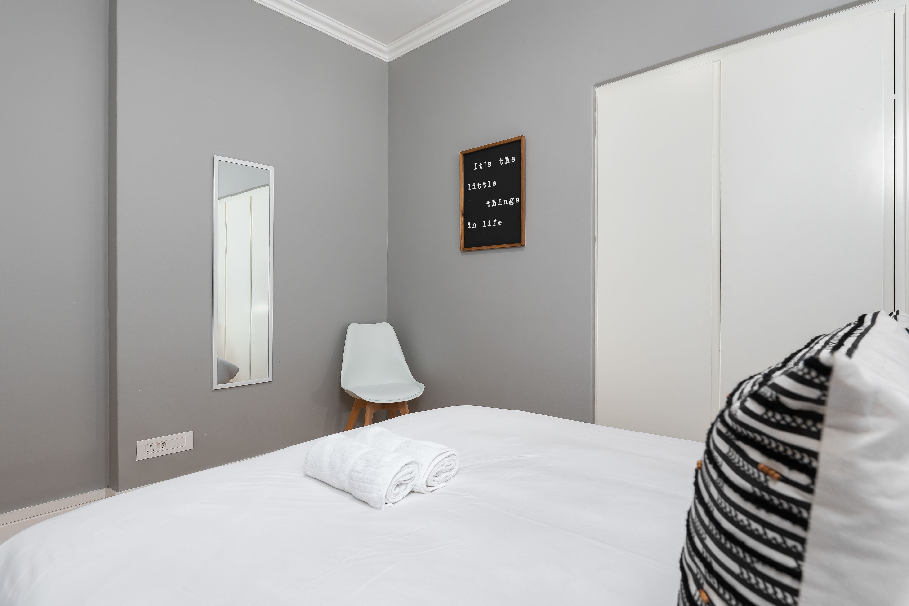 A neatly made bed with a rolled white towel, a mirror on the wall, a framed inspirational quote, and a white chair.