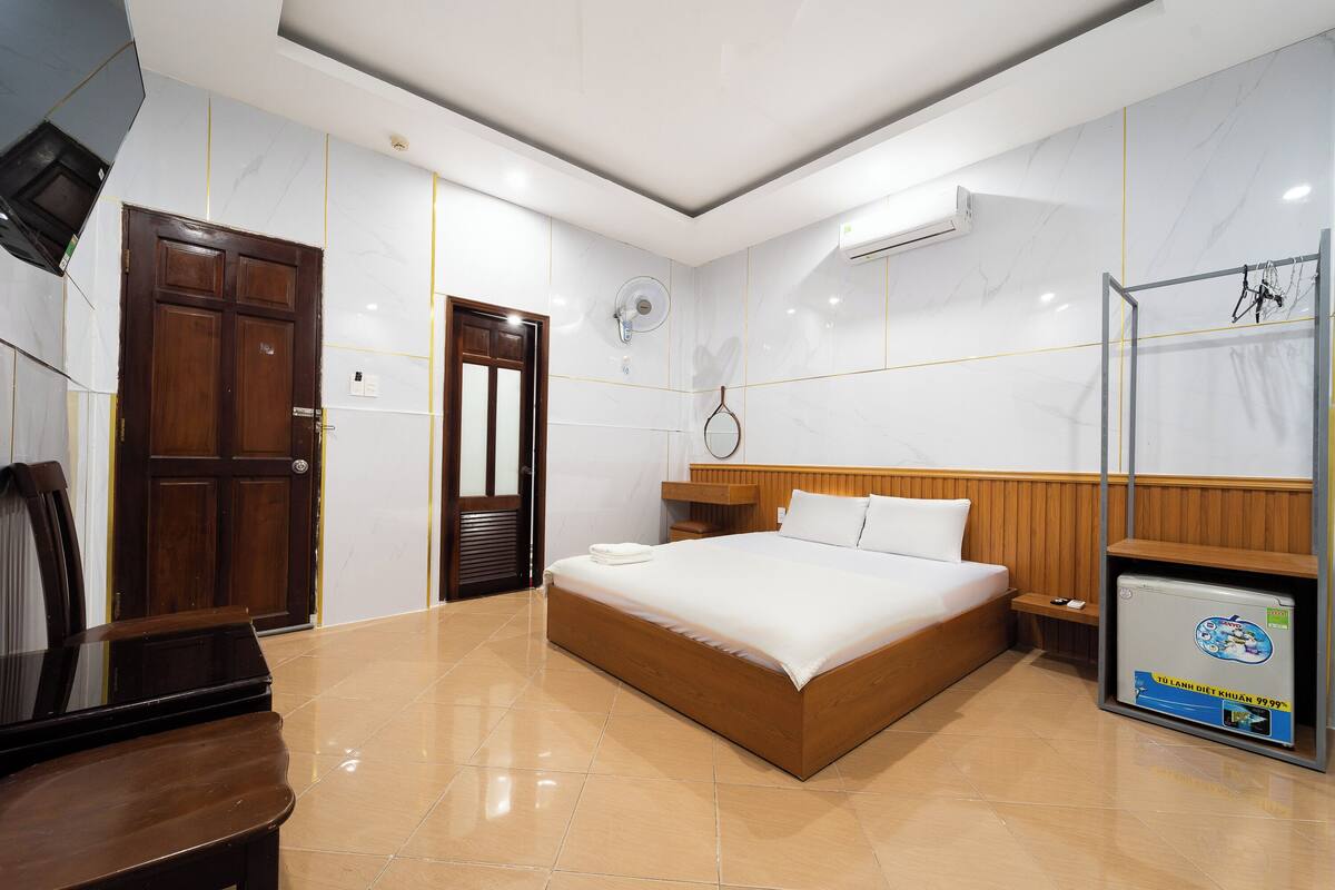 A modern hotel room with a large bed, wooden headboard, a small bedside table, a TV, and a built-in safe.