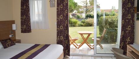 A hotel room with a bed, a small table, and a chair. There is a sliding glass door leading to a patio with a view of the garden.