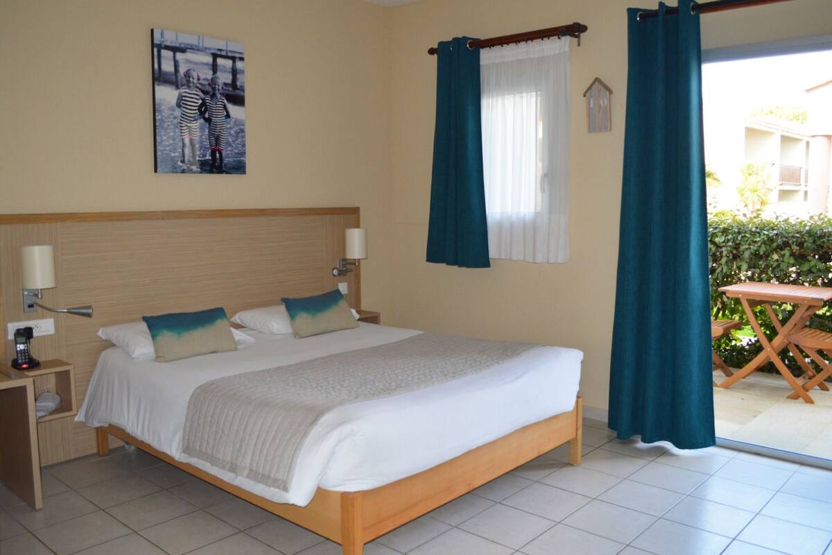 Quarto de hotel com uma cama grande, duas mesas de cabeceira, uma luminária de apoio e uma janela com cortinas, oferecendo vista para o exterior.