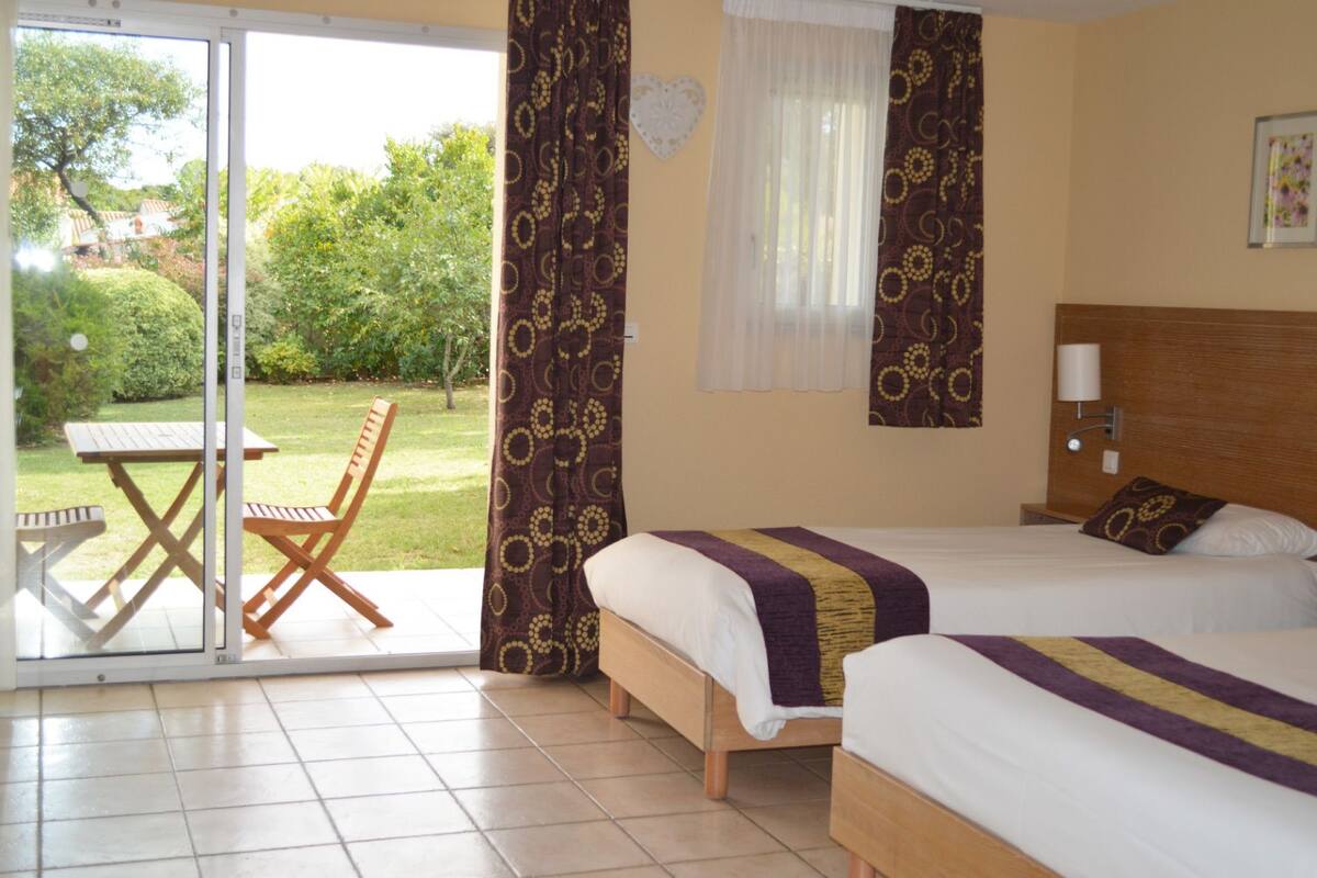 Quarto de hotel com duas camas, uma mesa e cadeiras no exterior, e vista para um jardim.