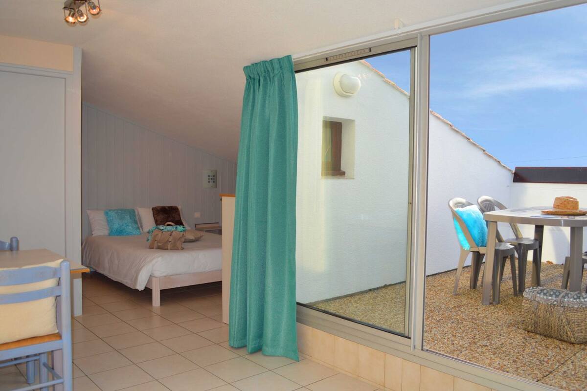 Quarto com cama, mesa e cadeira, varanda com mesa e cadeiras, e janela com cortina.