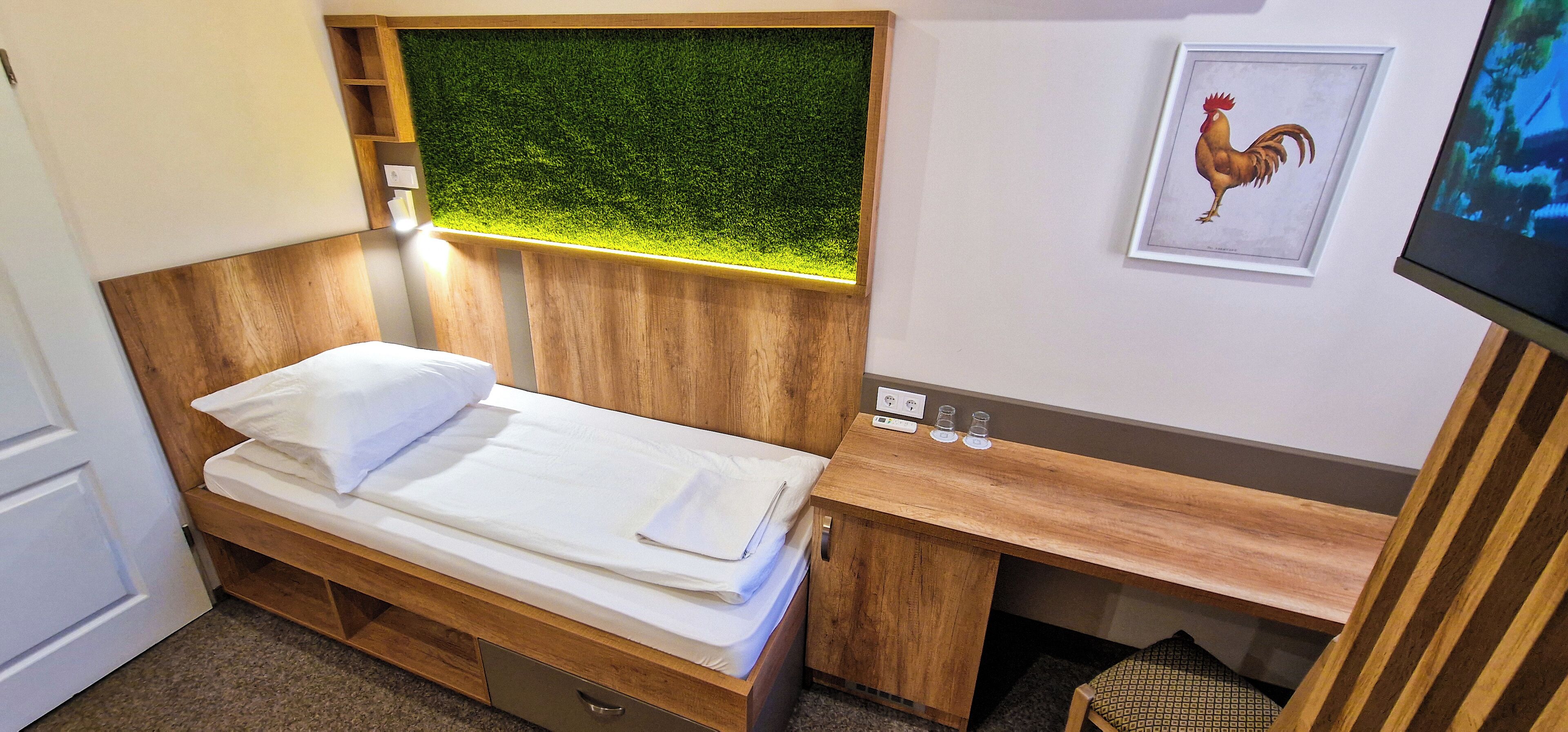 Une chambre à coucher de taille modeste, comprenant un lit, un bureau en bois, une petite étagère, un aquarium fixé au mur et un tableau représentant un coq.