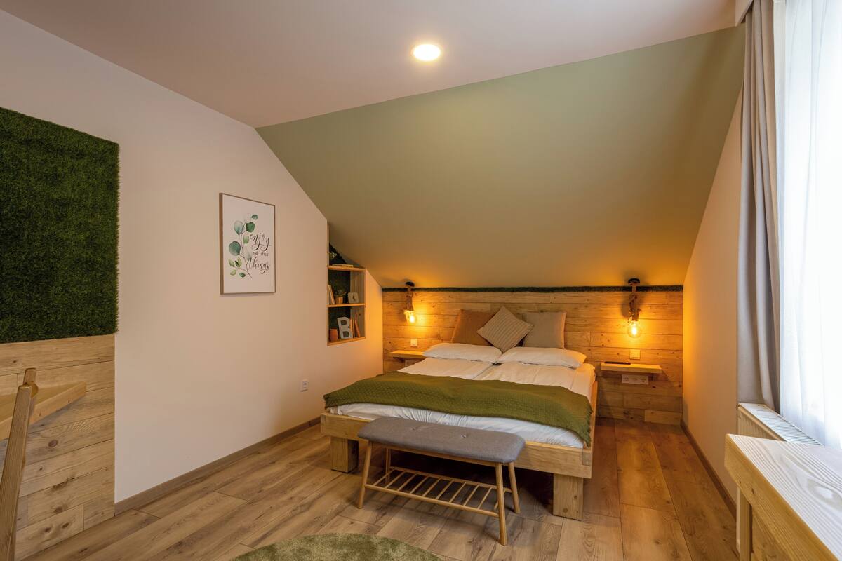 A bedroom with a sloped ceiling, wooden flooring, a bed with a green blanket, two bedside tables with wall-mounted lamps, and a wooden bench.