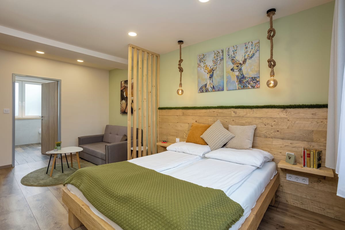 A bedroom with a wooden headboard, a bed with white linens, a green throw blanket, a small round table with a plant, and a chair. There are two paintings on the wall, a wooden partition, and a shelf with books and a small decorative item.