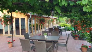 Un patio al aire libre con mobiliario de mimbre, piso de ladrillo y una pérgola de madera cubierta de flores rojas.