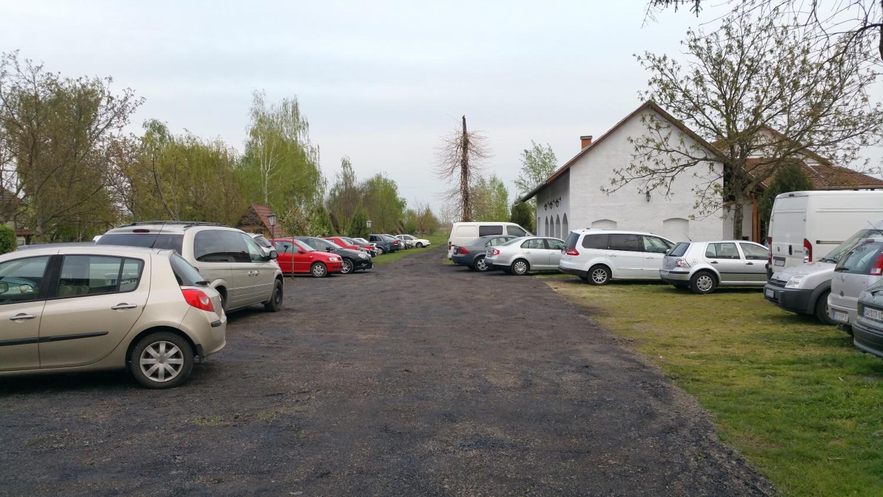 Un estacionamiento con varios autos estacionados a ambos lados, un edificio con techo inclinado y árboles al fondo.