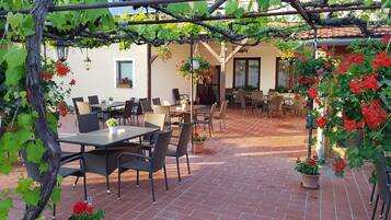 An outdoor patio area with tables and chairs, surrounded by potted plants and a pergola with vines.