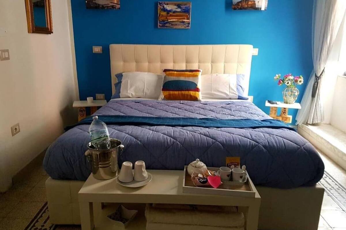 A bedroom with a blue wall, a white bed with a blue quilt, a bedside table with a water bottle, cups, and a tray with snacks.