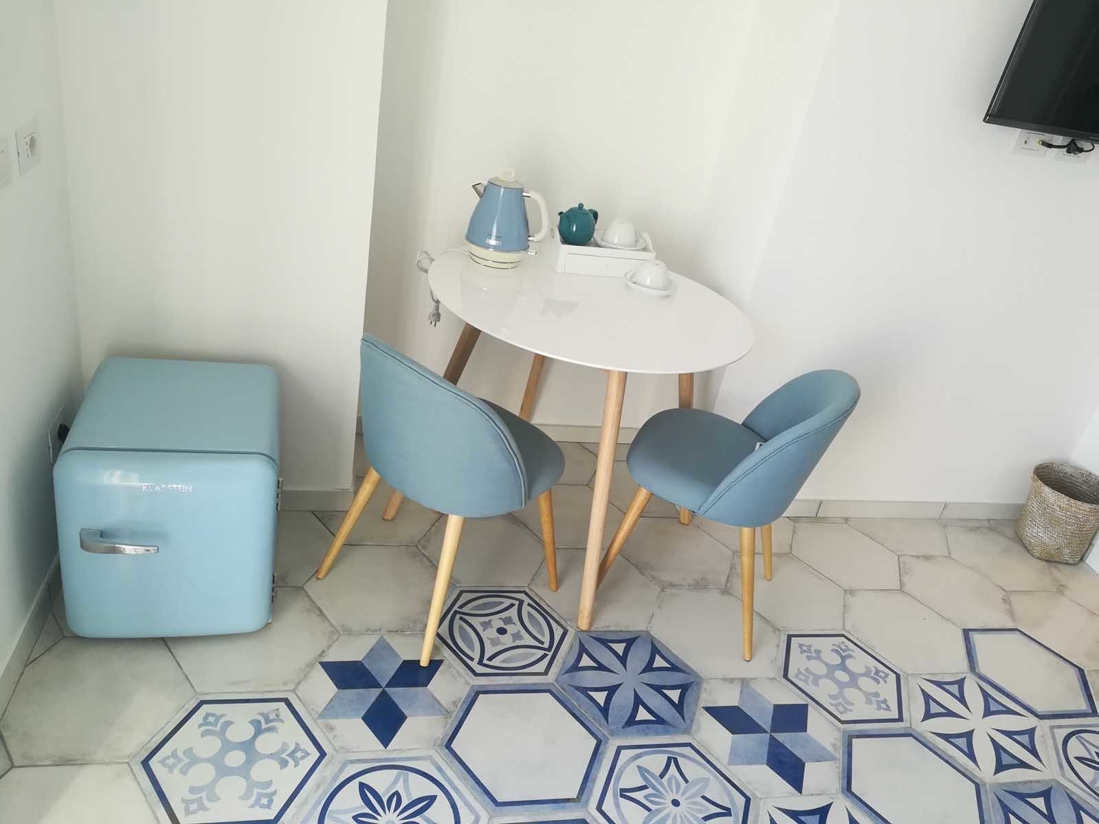 A small kitchenette with a blue appliance, a white table with two blue chairs, and a patterned floor.