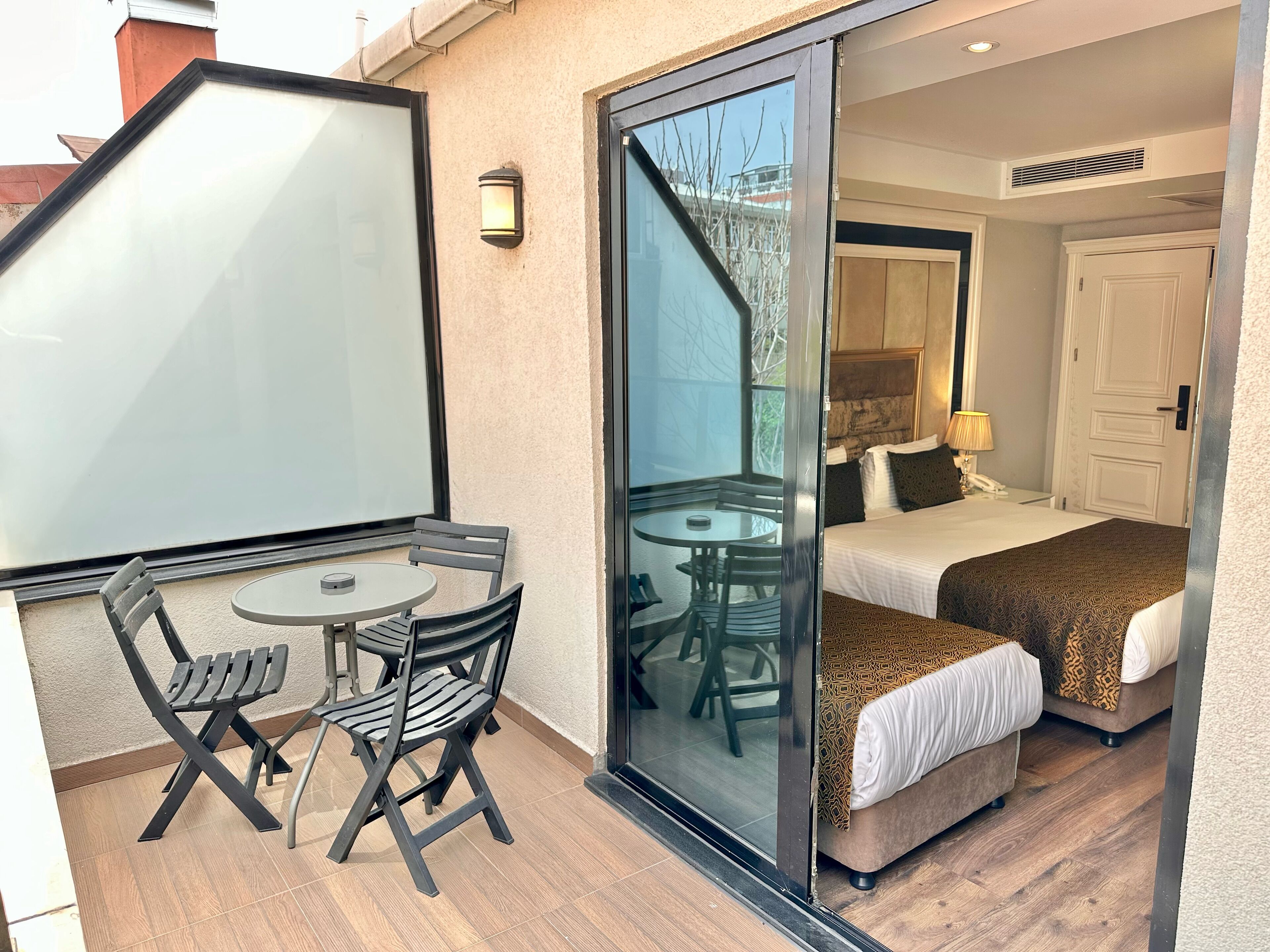 A hotel room with a glass door leading to a balcony, a bed with patterned bedding, a small round table with chairs, and a wall-mounted light fixture.