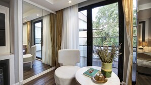 A modern hotel room with a large sliding glass door leading to a balcony, a white armchair, a small round table with a book and a vase of flowers, and a view of greenery outside.