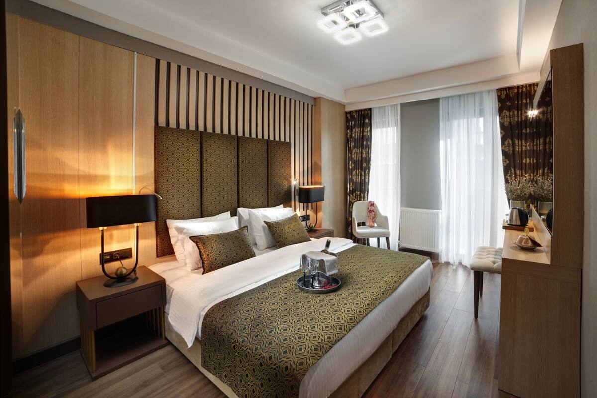 Quarto de hotel moderno com uma cama grande, uma mesinha de cabeceira com abajur, uma pequena mesa com chaleira e uma cadeira.