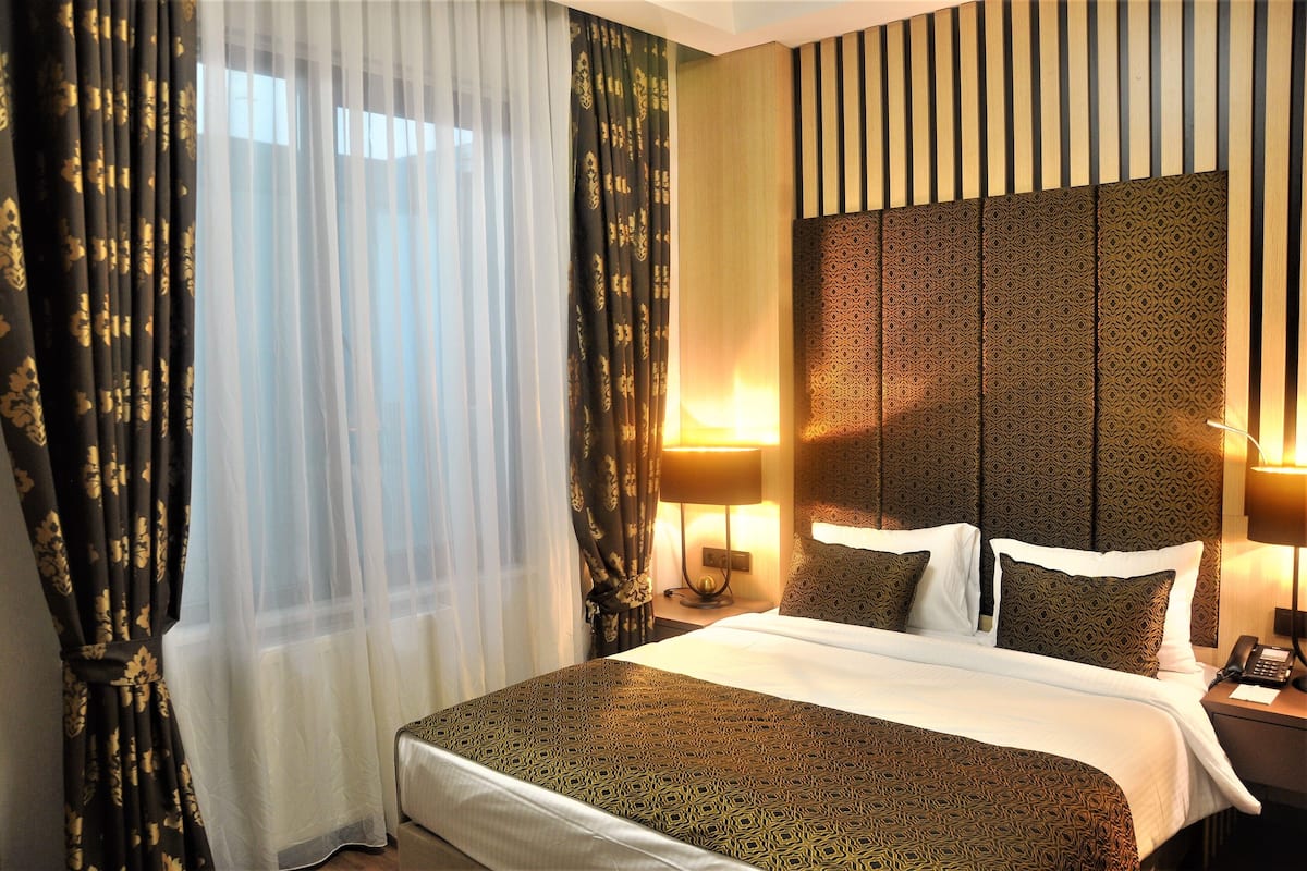 Quarto de hotel com uma cama grande, duas luminárias de cabeceira, um painel de cama estampado e cortinas finas com padrão floral.