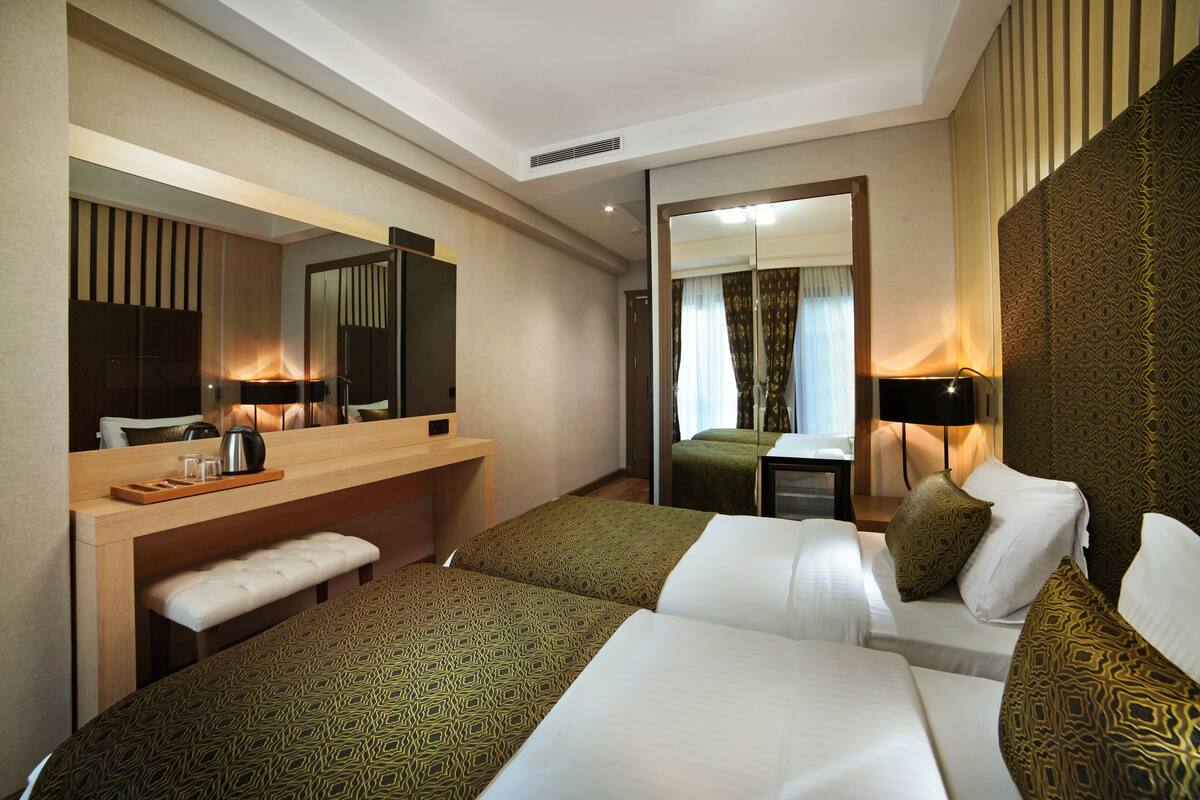 Quarto de hotel com duas camas, uma escrivaninha com cadeira, um banco, um espelho e parede estampada.