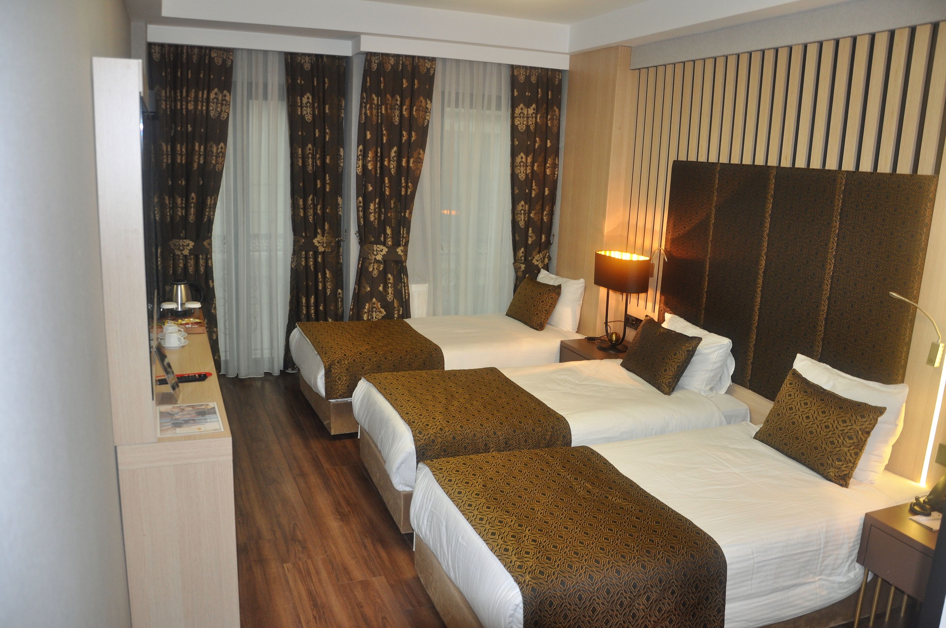 Habitación de hotel con dos camas, piso de madera, una ventana grande con cortinas y cabecera con listones verticales.