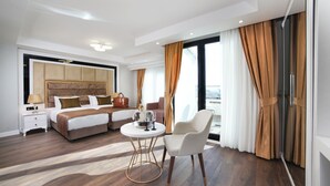 A modern hotel room with a large bed, a bedside table with a lamp, a small round table with a vase, a white armchair, and a view of the city through large windows.