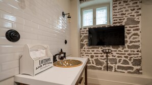 A bathroom with white tiled walls, a stone accent wall, a mounted TV, a sink with a decorative bowl, and a tissue box labeled "Lotions and Potions". - 29A Guesthouse (Plovdiv)