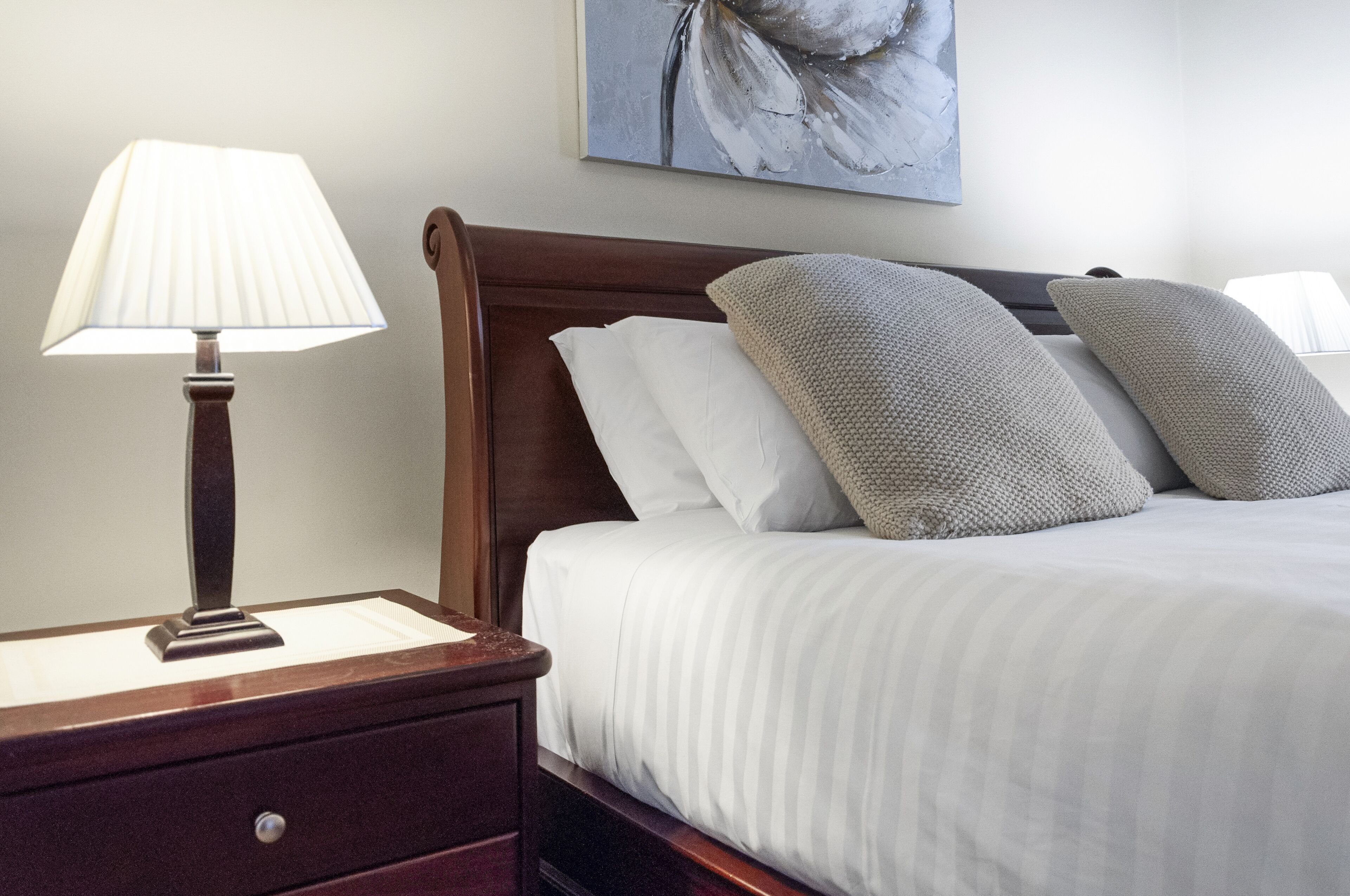A neatly made bed with white linens and two pillows, a wooden nightstand with a lamp, and a painting on the wall.