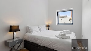 A modern bedroom with a bed, two bedside tables, a window, and a view of buildings outside. - City Centre Spacious Apartment (Exeter)