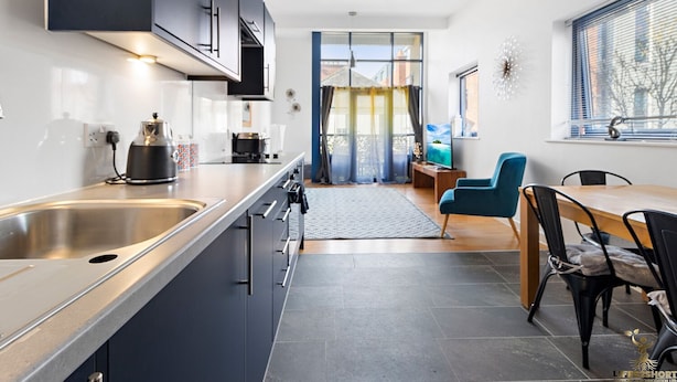 A modern kitchen with a central island, built-in appliances, and a dining area with a blue chair and wooden table. - City Centre Spacious Apartment (Exeter)