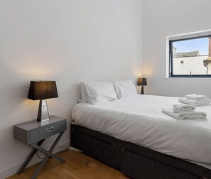 Ein modernes Schlafzimmer mit einem Bett, zwei Nachttischen, einem Fenster und Blick auf Gebäude.