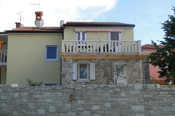 A two-story building with a stone facade, a balcony with white railings, and a small window with shutters.