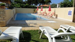 Ein Swimmingpool mit Liegestühlen und Sonnenschirmen, umgeben von einer niedrigen Steinmauer und Grünpflanzen.