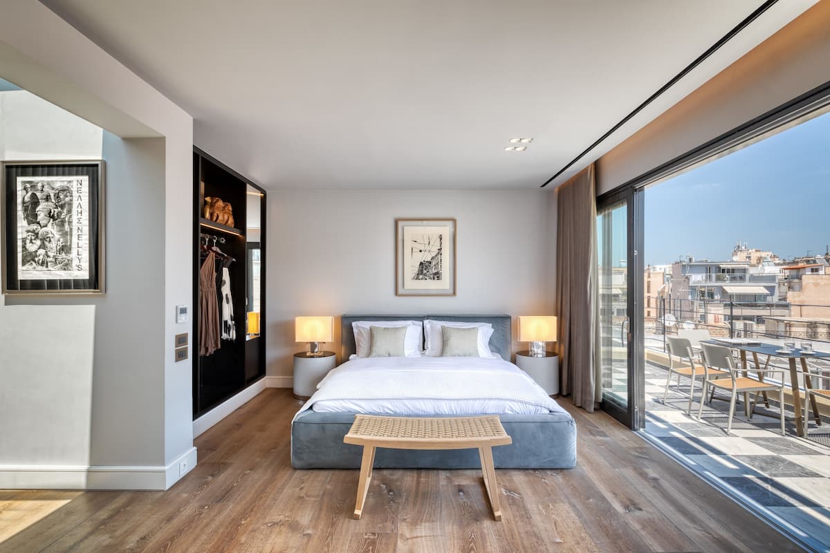Quarto moderno com uma cama grande, luminárias de cabeceira, uma mesa pequena e vista para a cidade por grandes janelas.