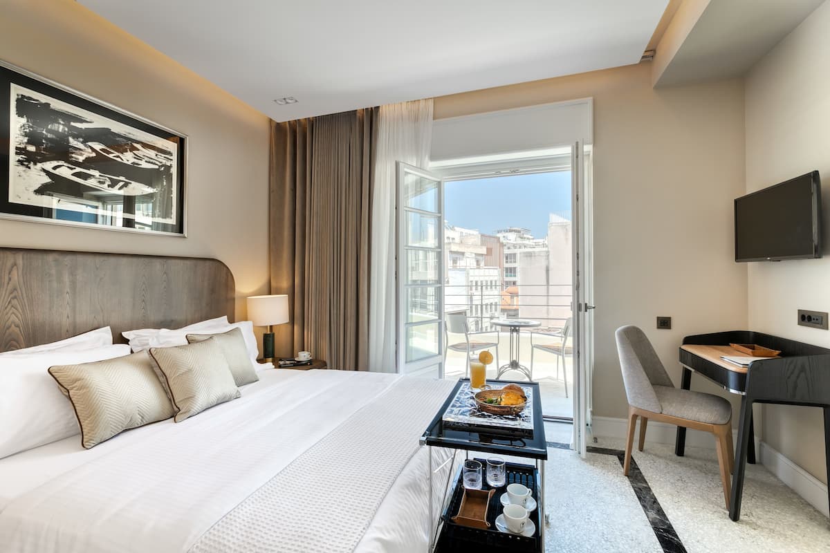 Quarto de hotel com uma cama grande, uma escrivaninha com cadeira, televisão e varanda com vista para a cidade.