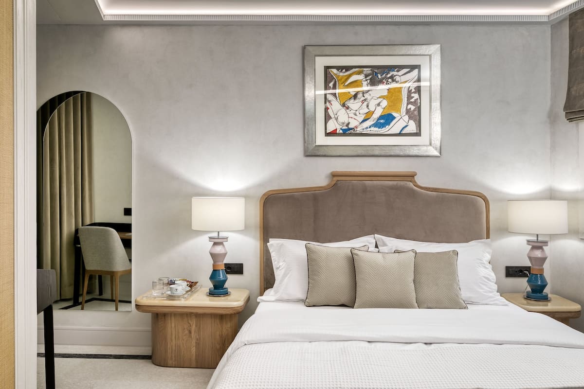 Quarto de hotel com uma cama grande, duas mesas de cabeceira com luminárias, uma obra de arte emoldurada na parede e um espelho na porta.
